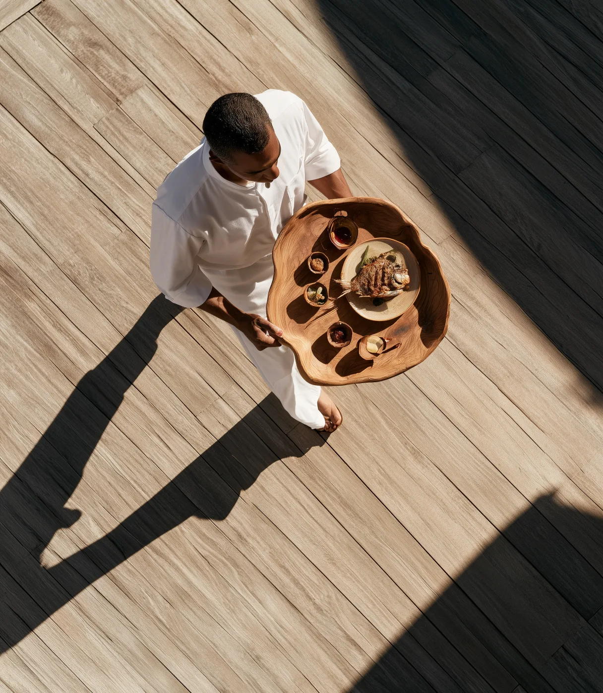 A host in white carrying a wooden tray of small dishes across a sunlit deck, seen from above