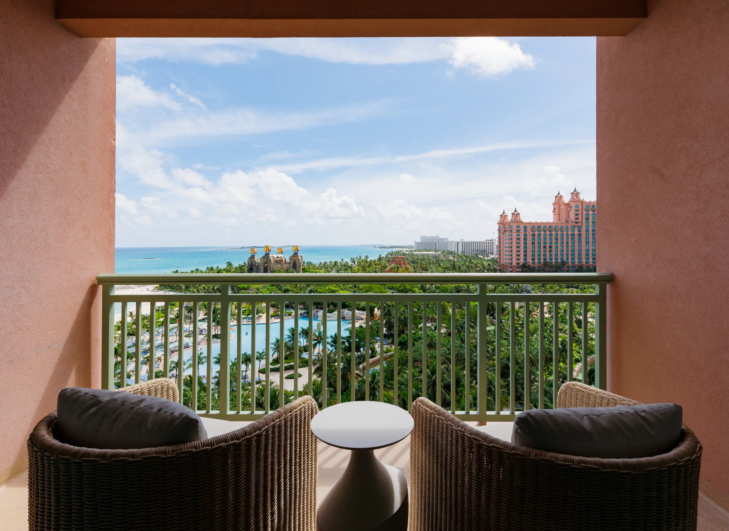 A suite balcony with two wicker armchairs looking over palms to the ocean and the Royal towers
