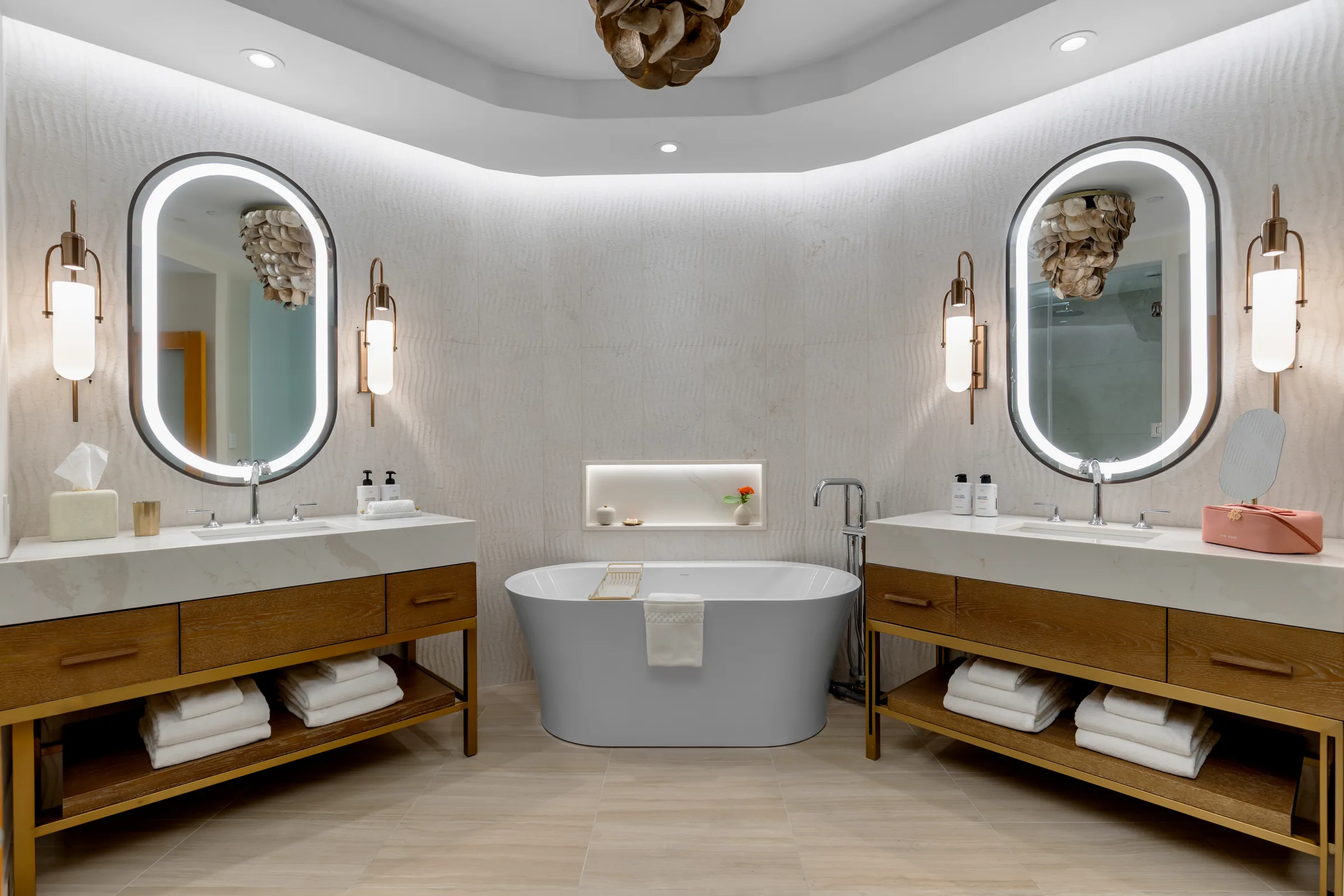 A suite bathroom: freestanding tub between two vanities with backlit oval mirrors